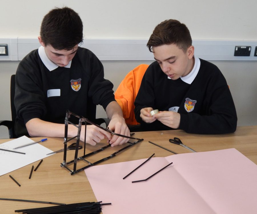 2 boys use straws to build a bridge on a desk