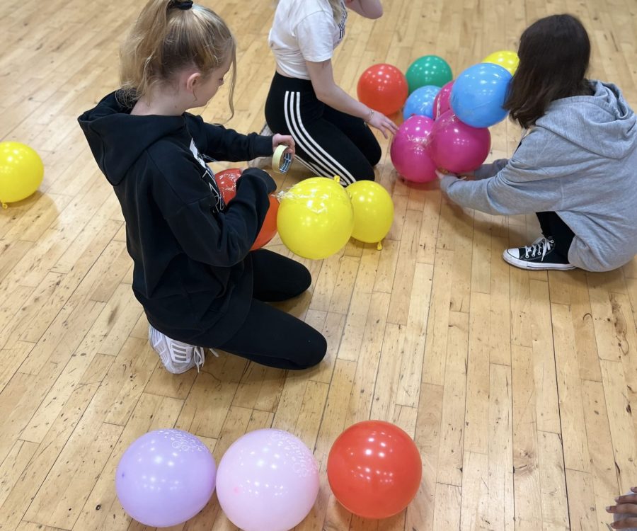 3 girls sit on a sports hall floor blowing up balloons